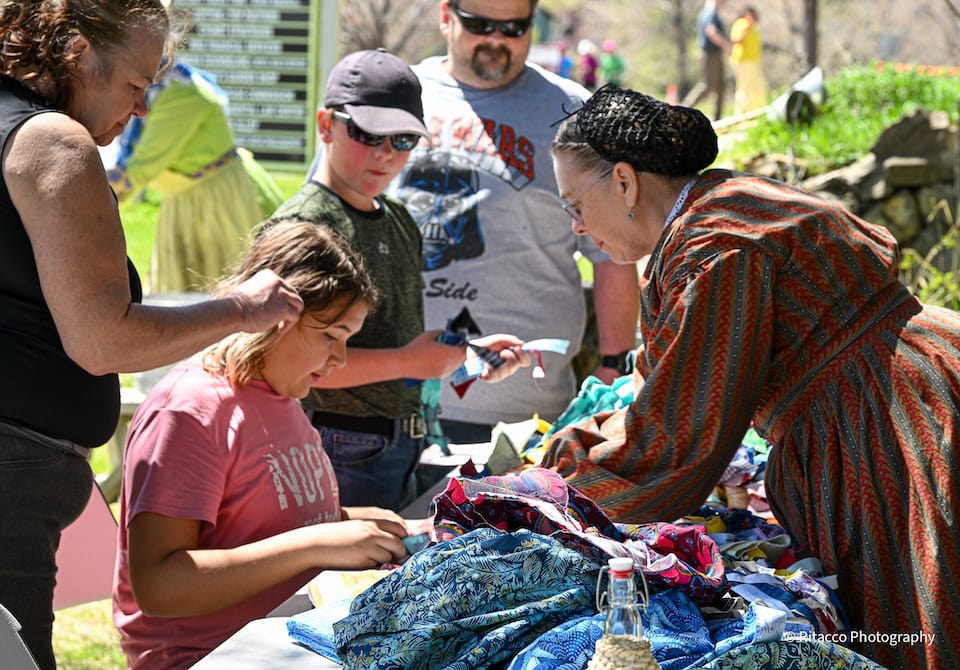 Woman and two children working on a project on a table outdoors with onlookers around.