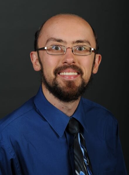 bald man with beard in a blue shirt and tie with glasses smiling